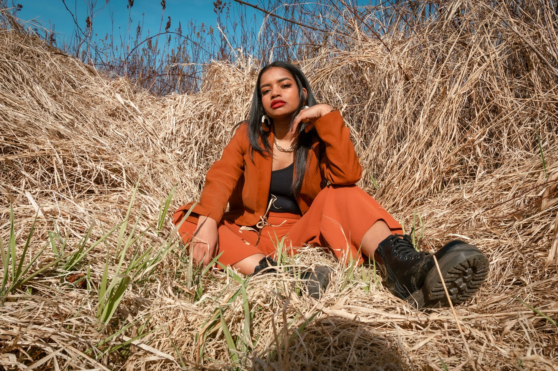Stylish woman in brown outfit poses elegantly in a field of dry grass under the bright sun.