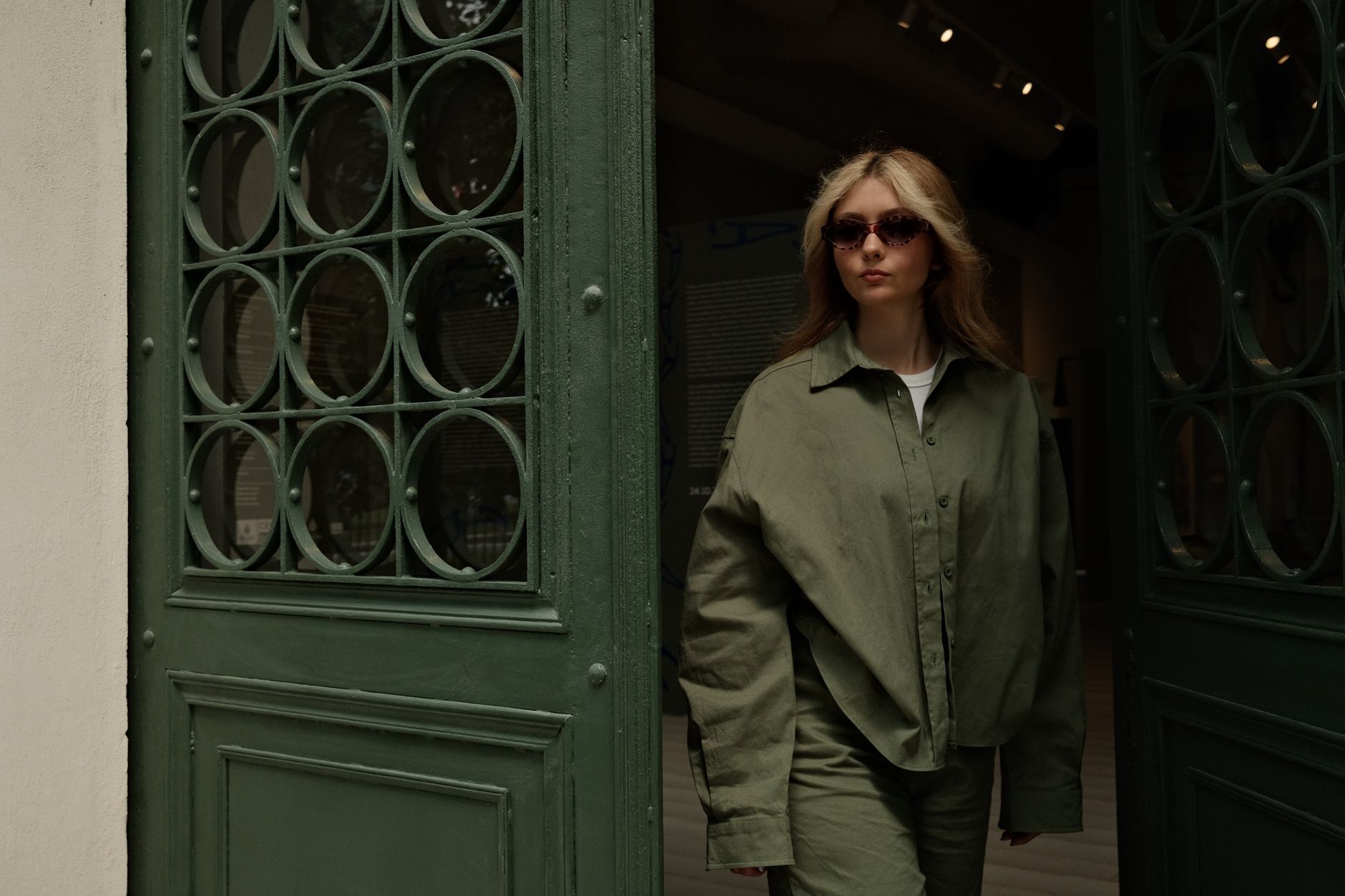 Woman in olive green oversized outfit and sunglasses exiting a vintage doorway.