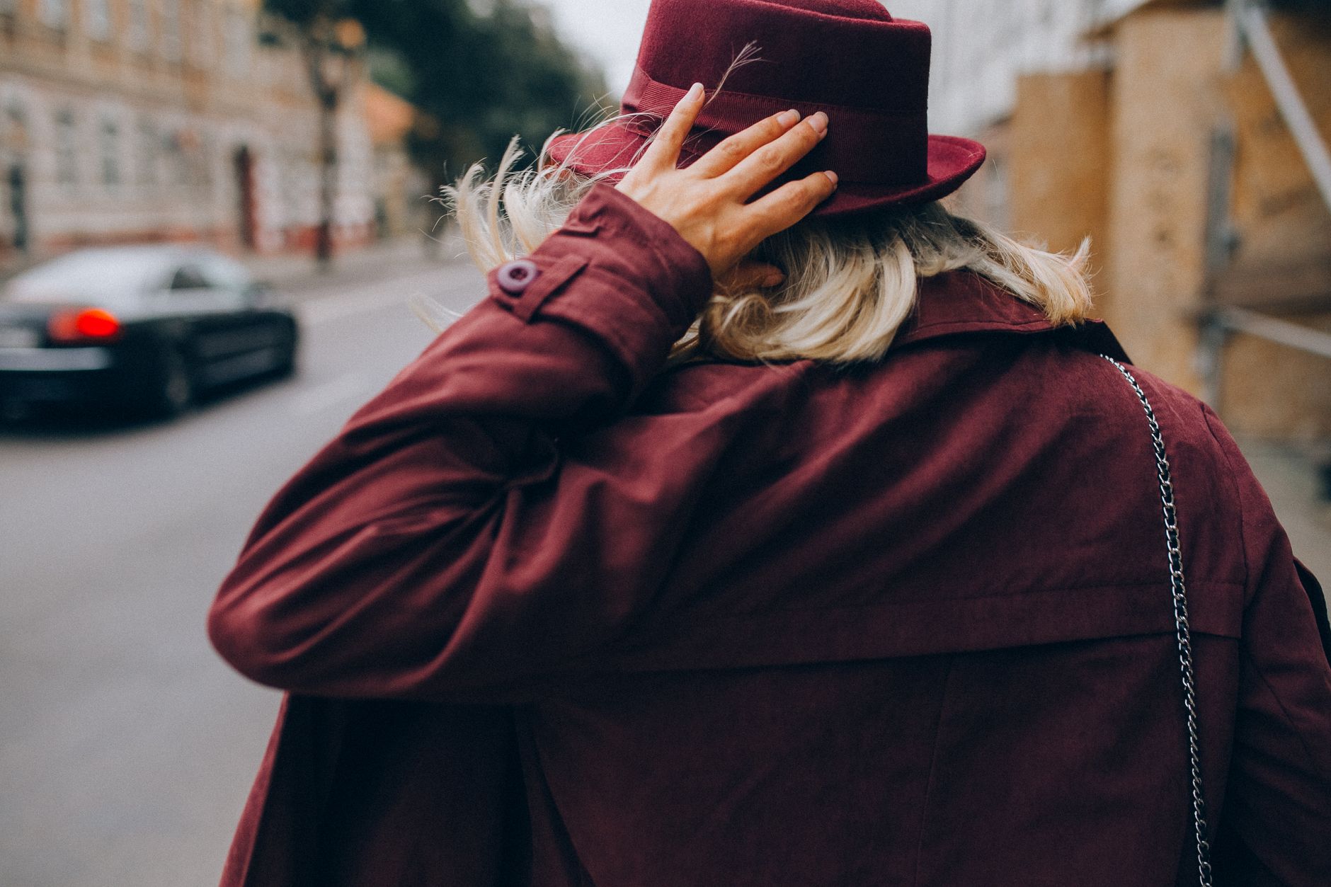 Stylish woman in burgundy outfit walks down a city street, hand on hat, with blurred car passing by.