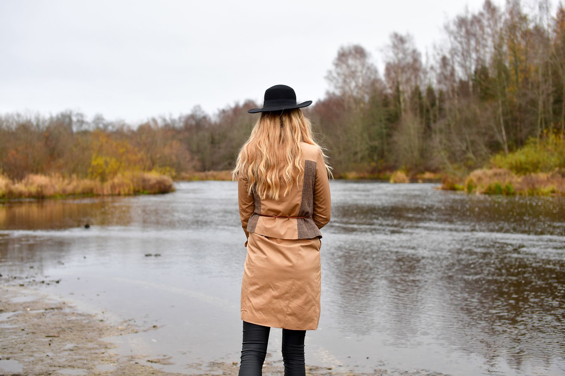 A woman in a trench coat and hat stands by a serene river in Jägala-Joa, creating a moody autumn scene.
