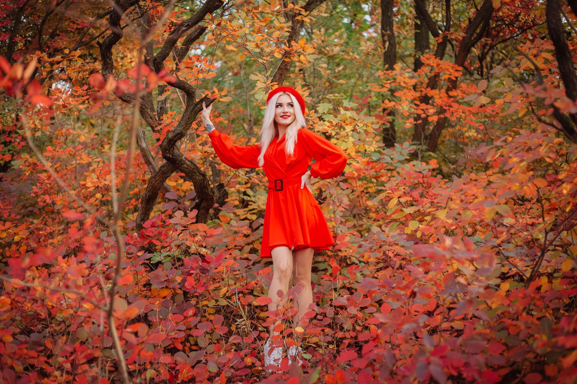 Blonde woman in a striking red dress stands among vibrant fall foliage in a forest.