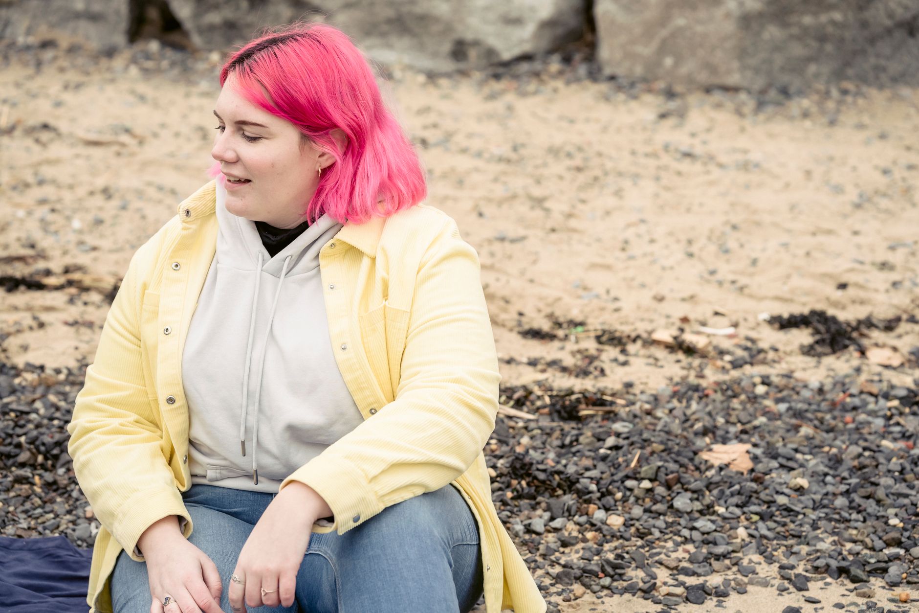 Positive plus size female with dyed hair wearing outerwear and jeans sitting on pebbled shore while spending time on waterfront