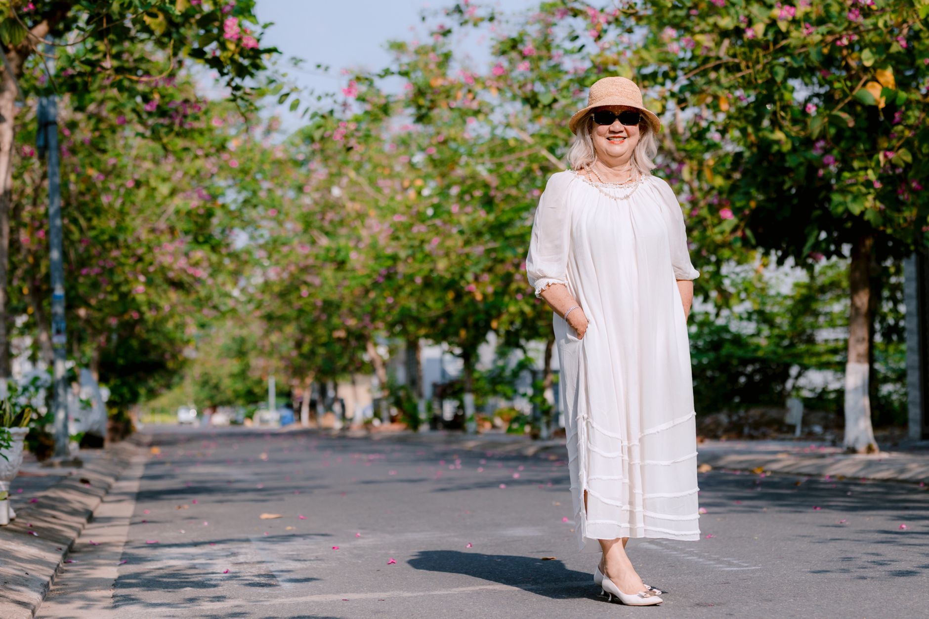 Senior woman in white dress and hat poses elegantly outdoors on a sunny day.