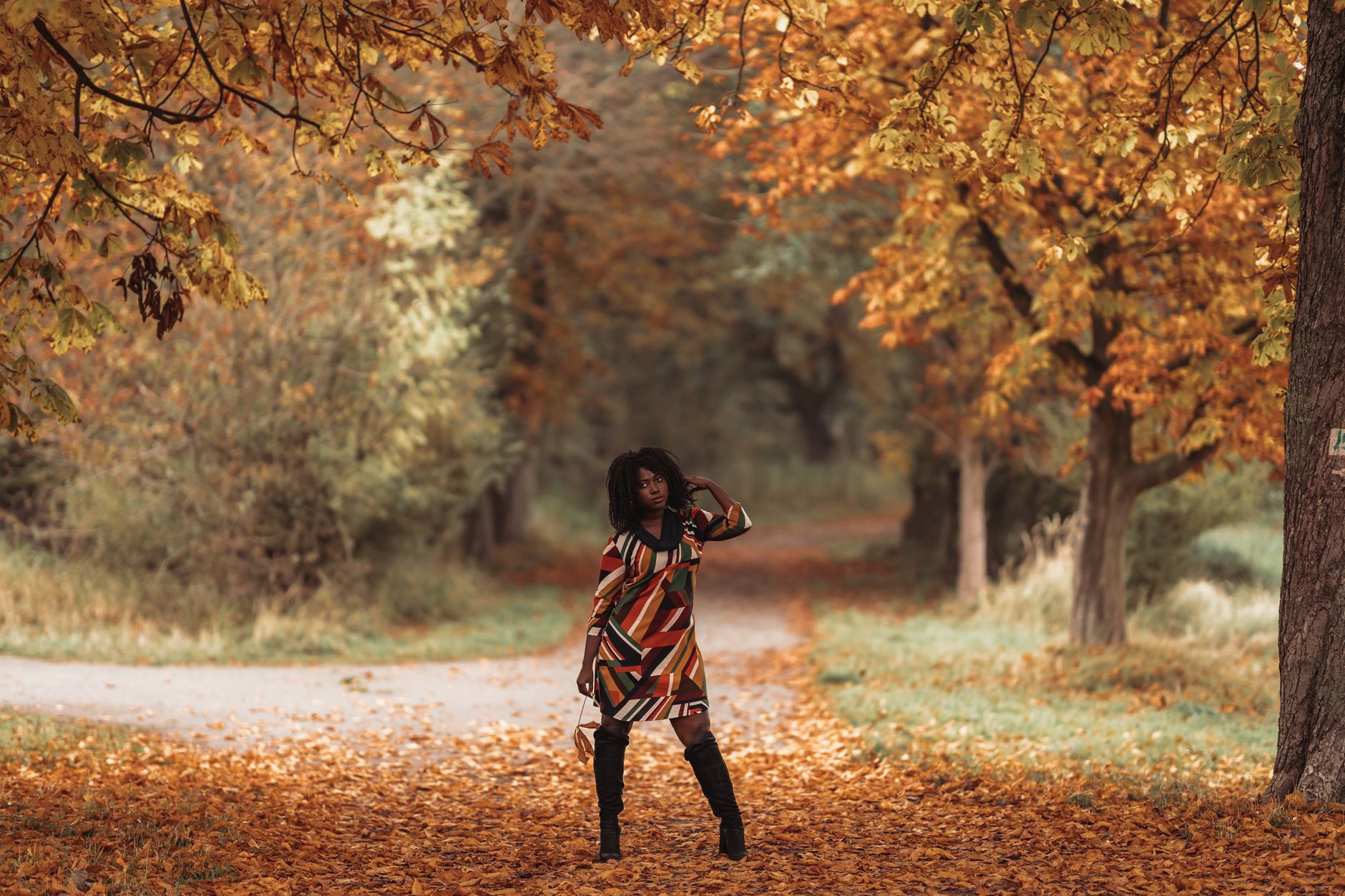 Stylish woman in vibrant dress and boots posing in a colorful autumn park.