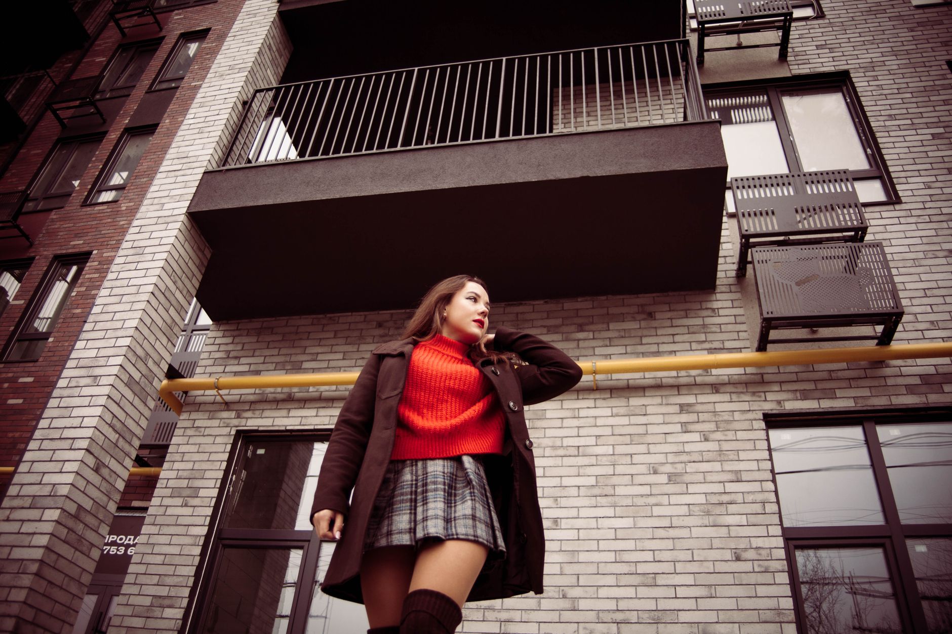 Stylish woman in red sweater and coat posing on a city street with modern architecture.