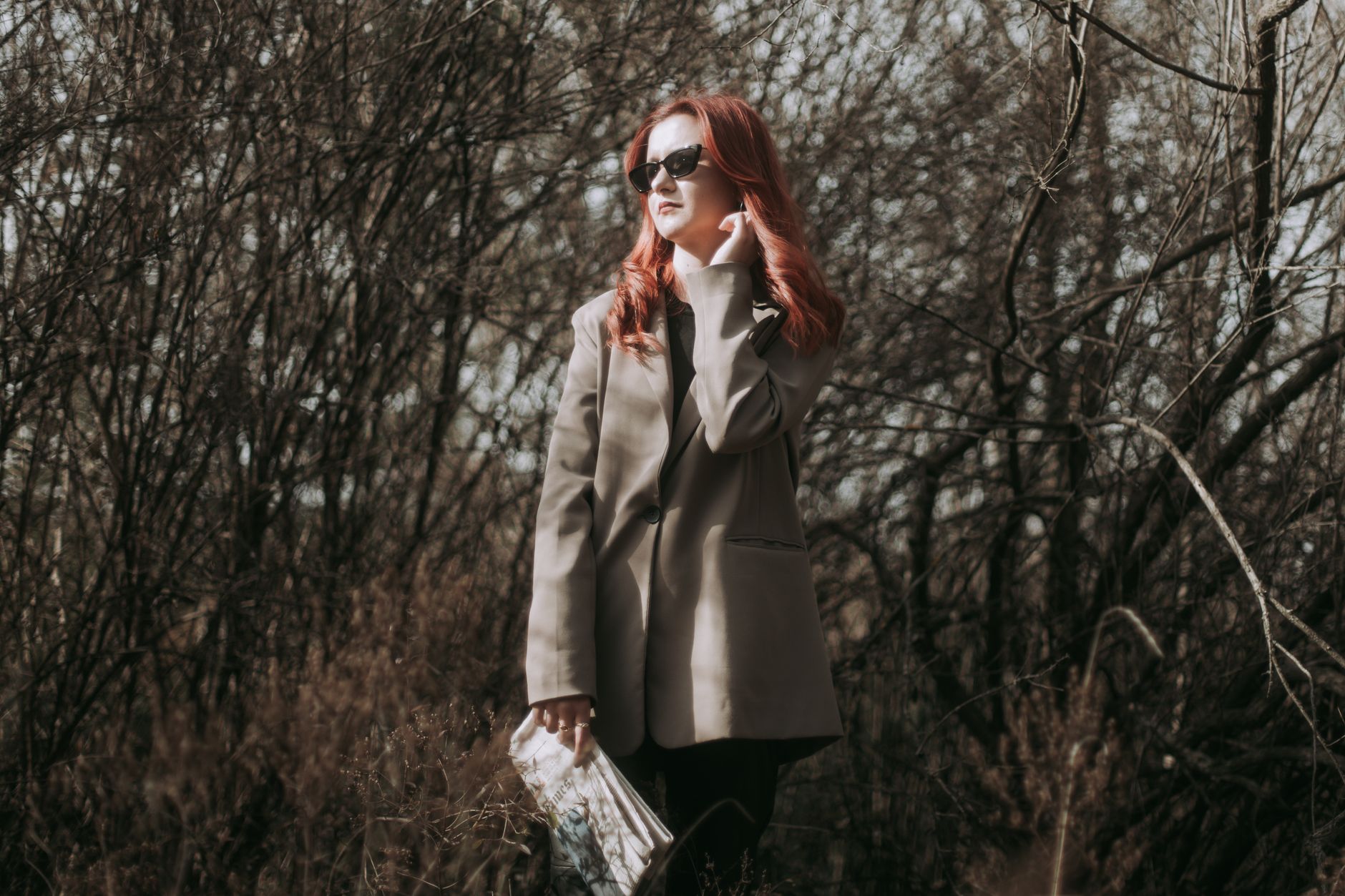 Redhead woman in gray blazer posing with a newspaper in an autumn forest setting.