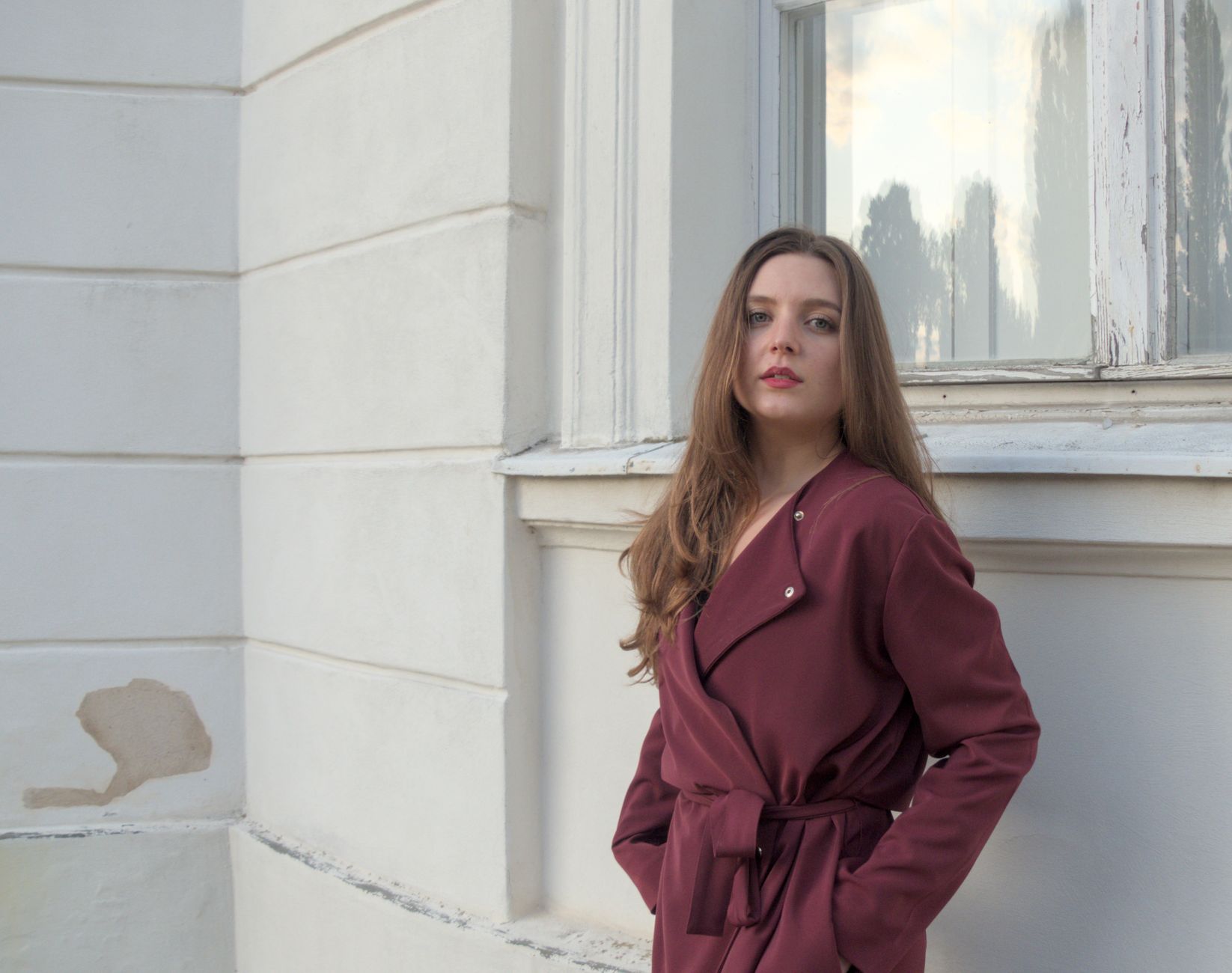 Fashion portrait of a young woman in a burgundy coat posing outdoors with an urban backdrop.