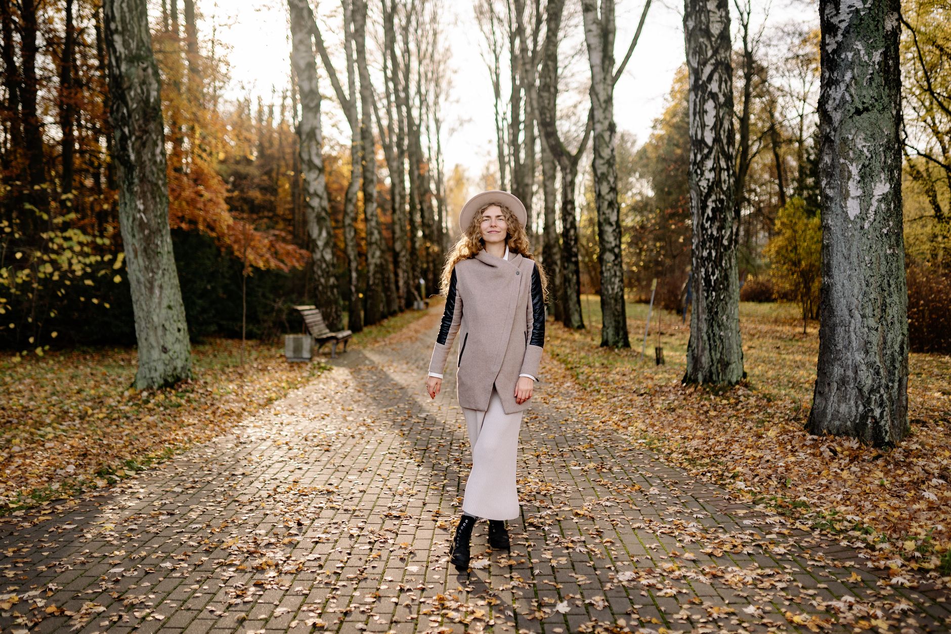 Woman in stylish coat and hat walks along a leaf-covered path in a sunny autumn park.