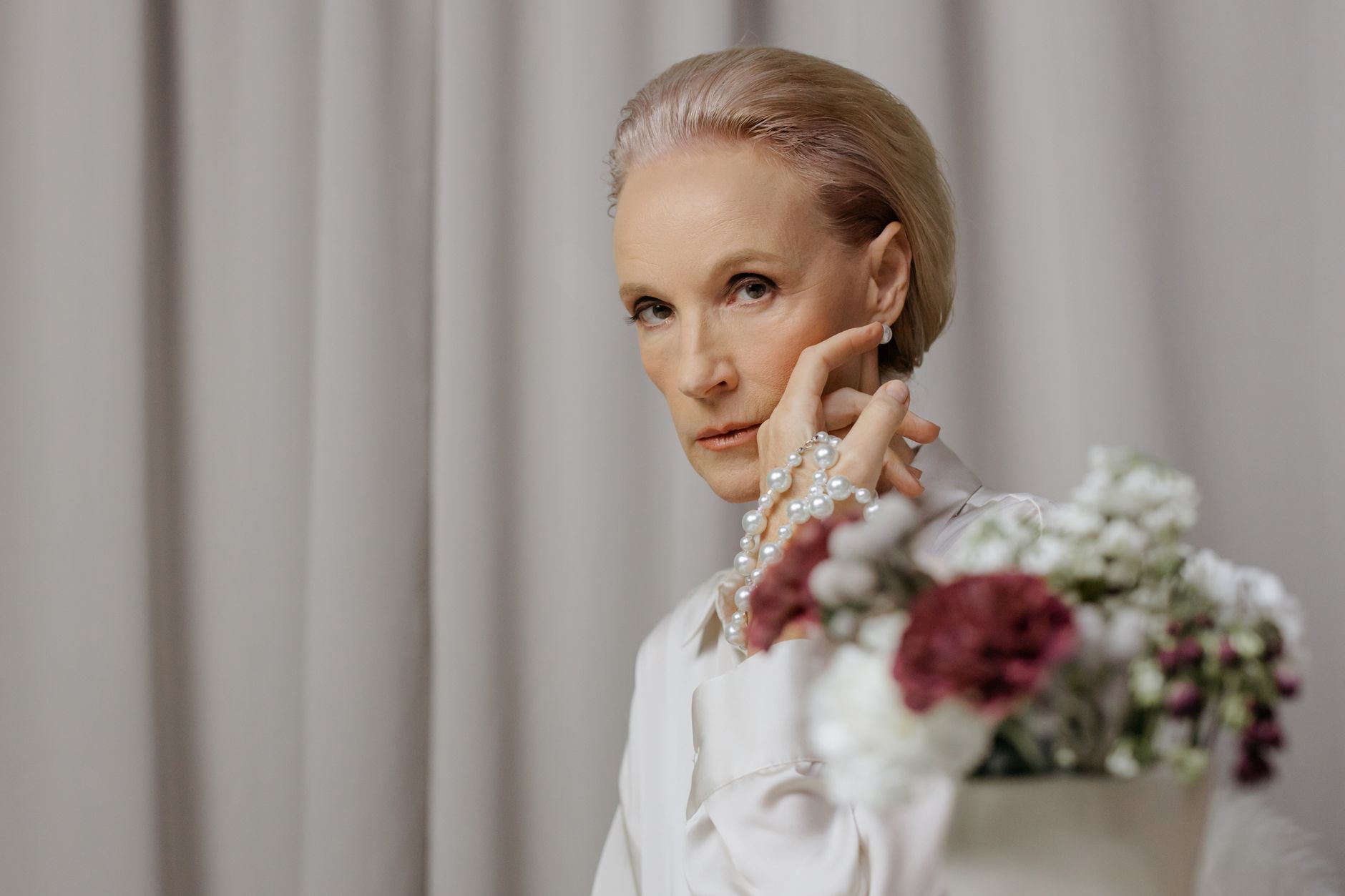 Elegant senior woman adorned with pearls and floral arrangement indoors.