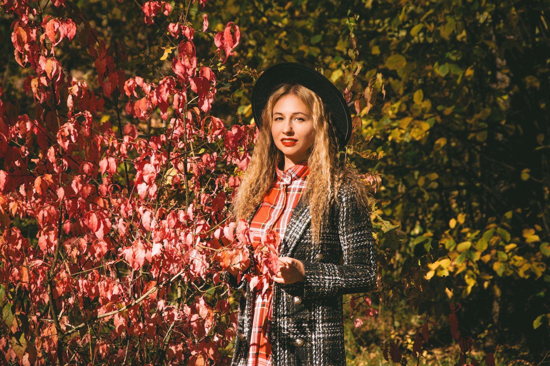 Stylish woman with curly hair in fall attire standing beside vibrant red foliage.