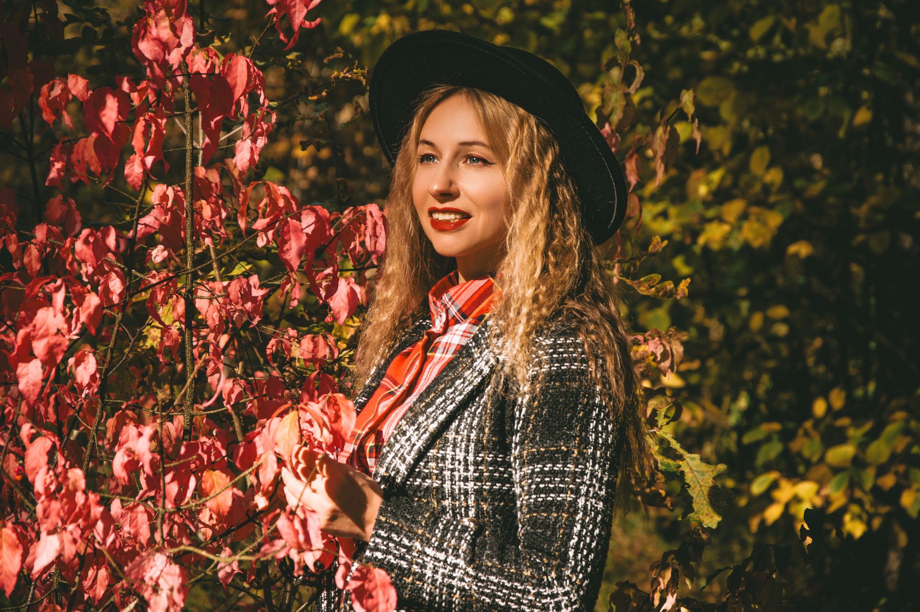 Stylish woman wearing a hat and plaid coat, surrounded by autumn foliage.