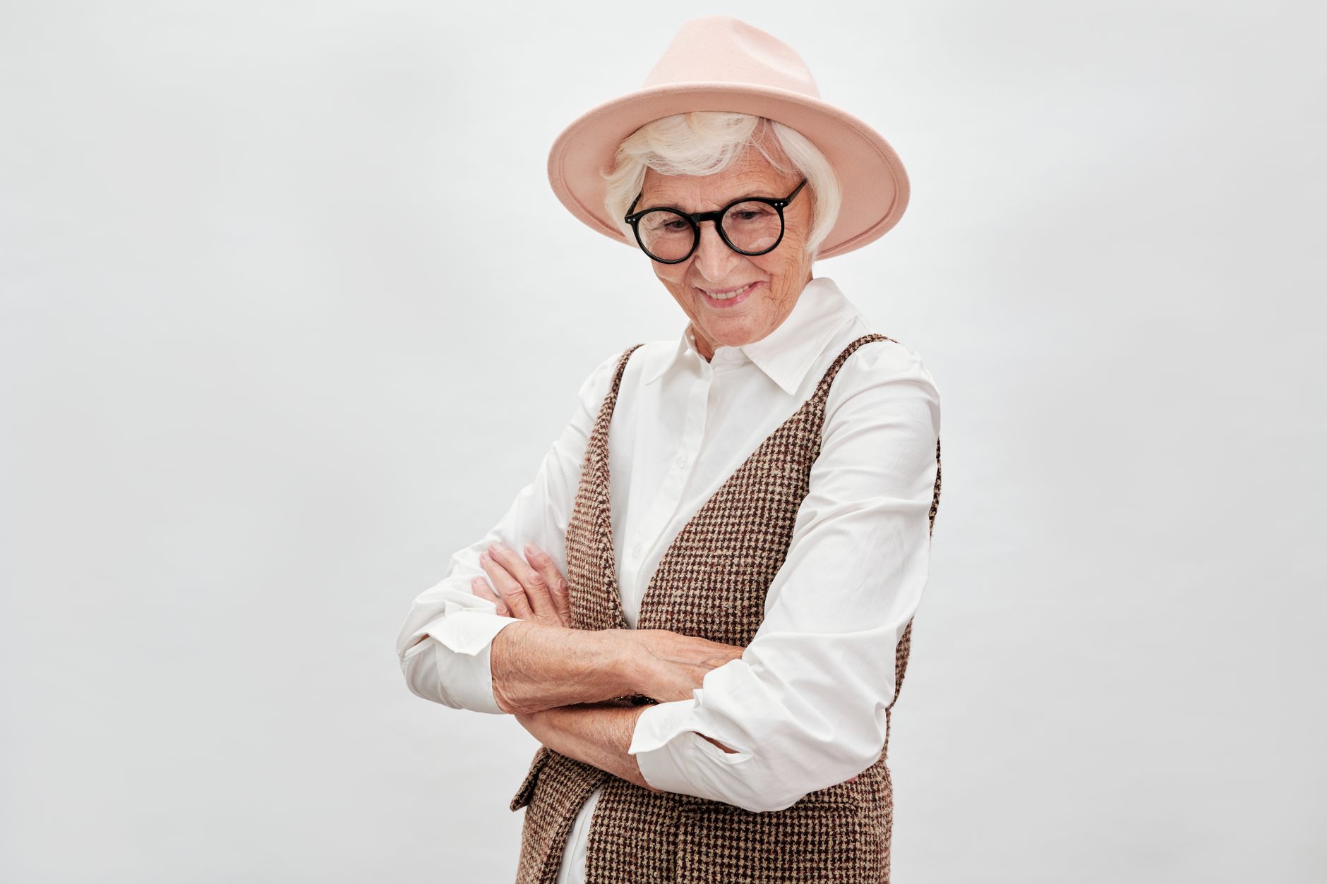 Elegant senior woman with glasses and hat, confidently posing with arms crossed.