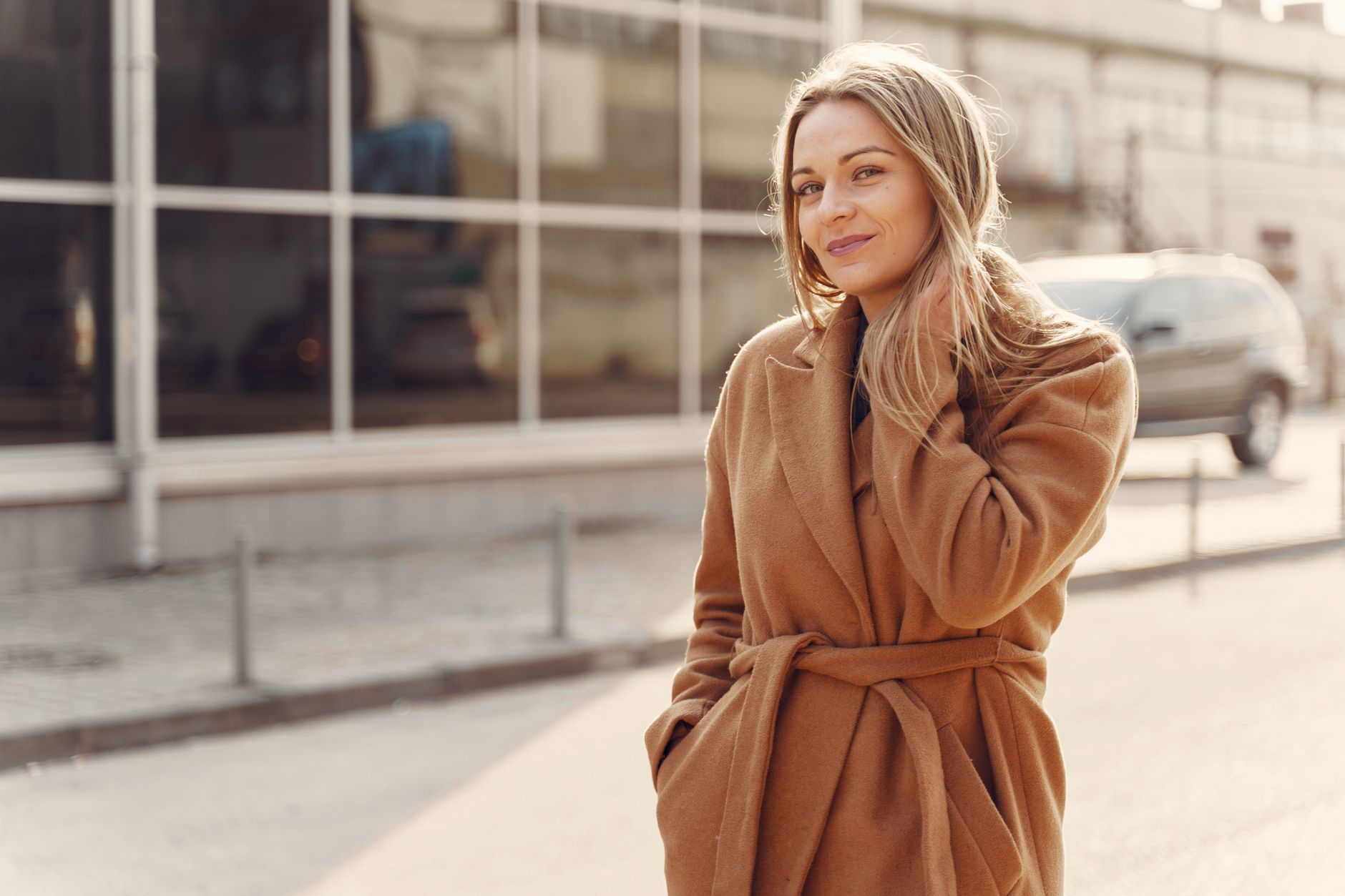 Positive female in stylish warm coat standing on city street against modern glass building and touching hair in back lit