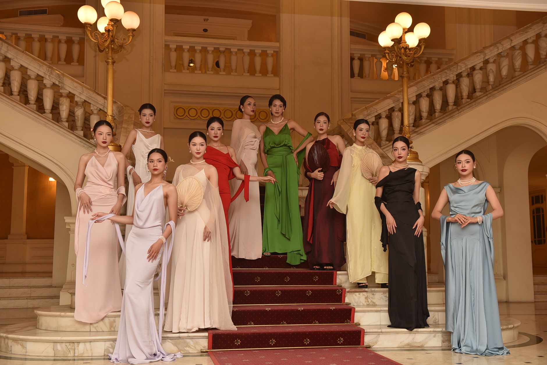 Group of diverse fashion models posing in elegant evening gowns on luxurious staircase indoors.