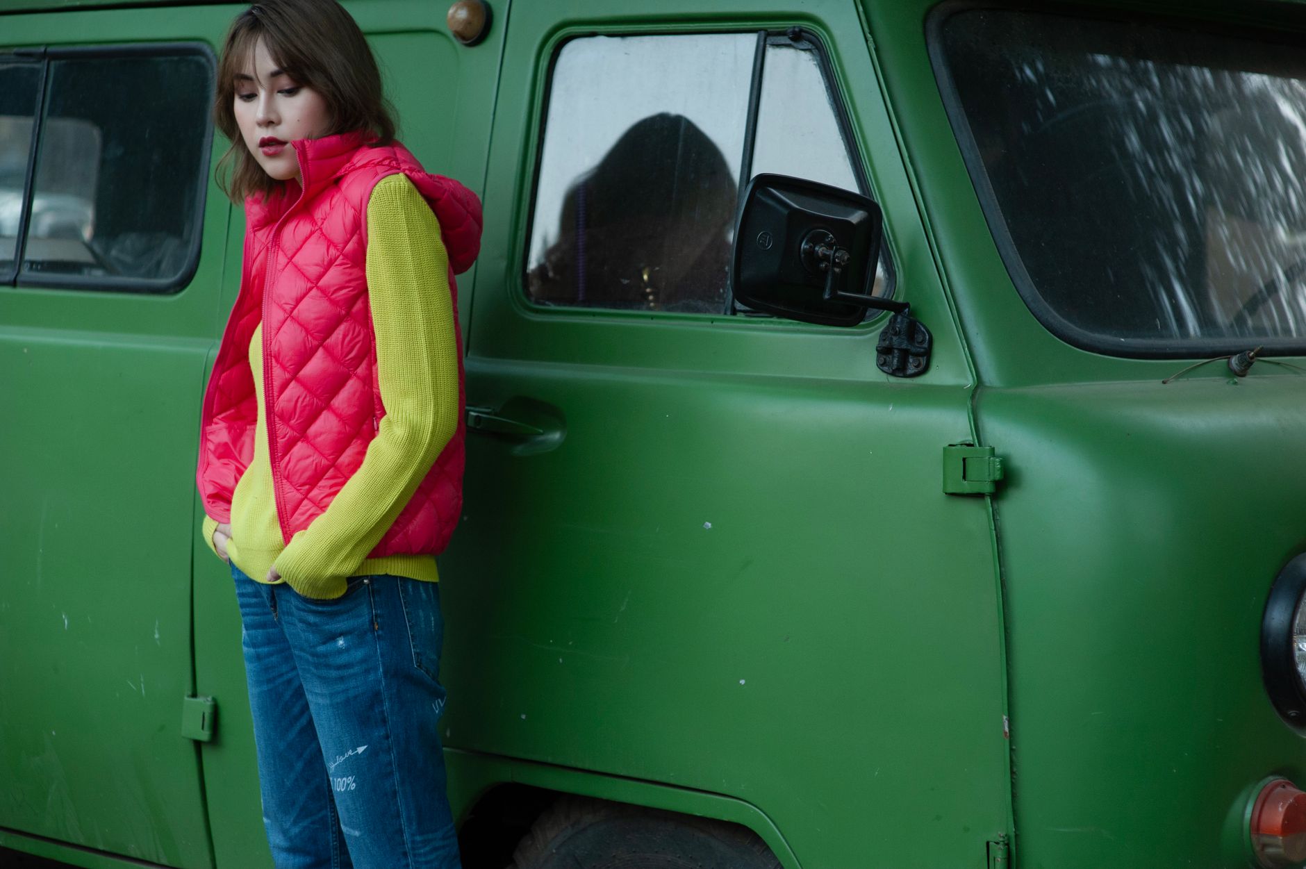 Fashionable woman wearing a red quilted vest and yellow sweater stands beside a retro green van.