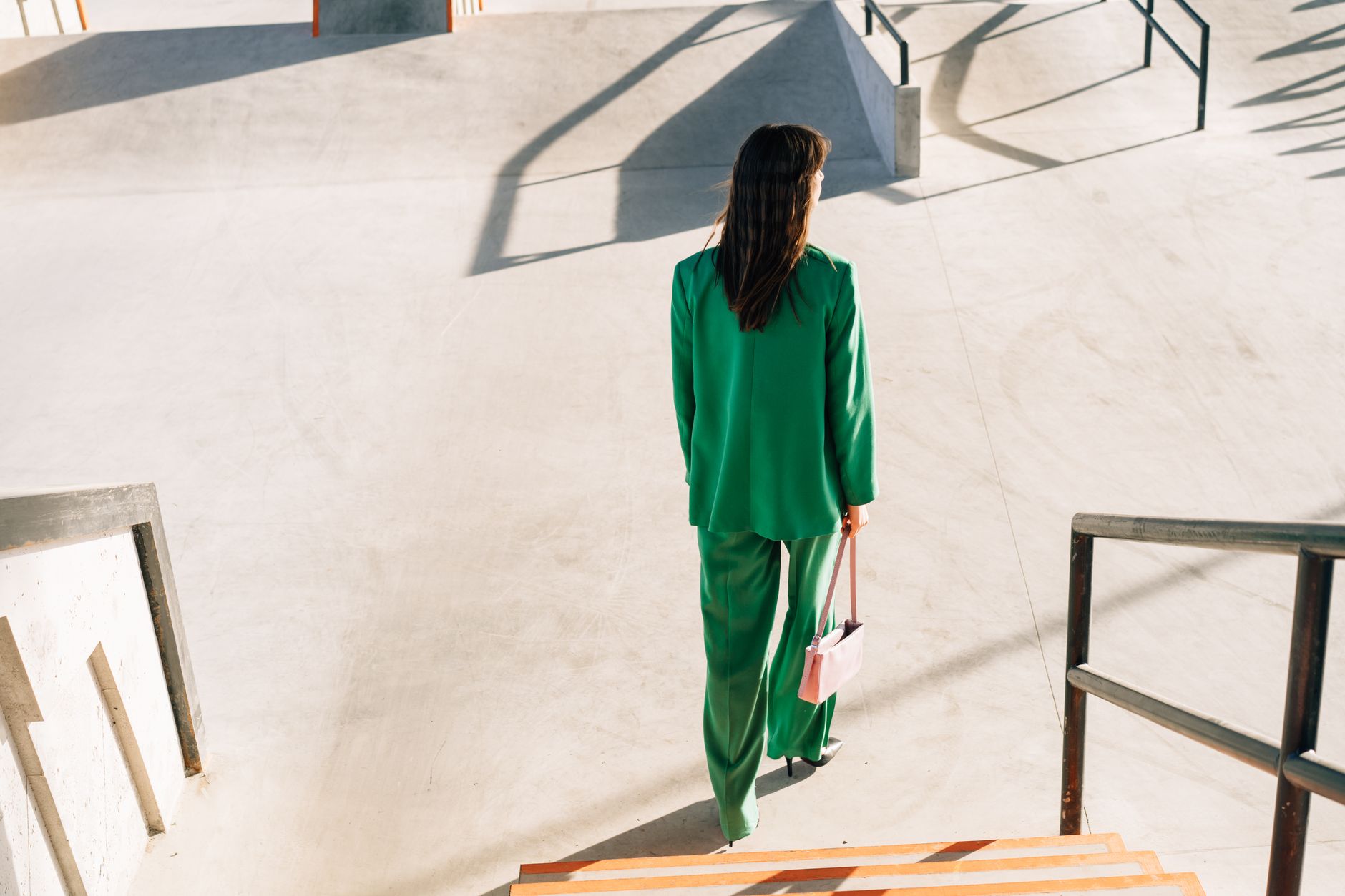 Fashionable woman in green suit climbs stairs carrying a pink handbag at sunset.