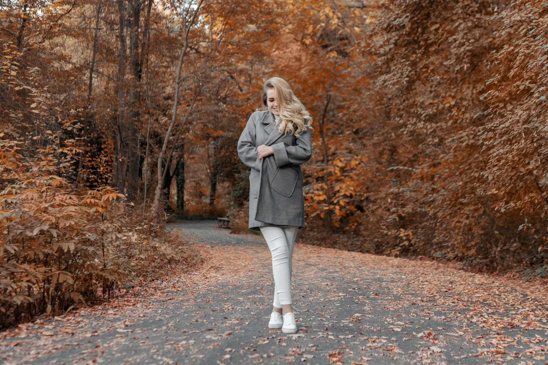 Full body happy young female bundling up in warm cozy coat while standing on asphalt road leading through autumn forest