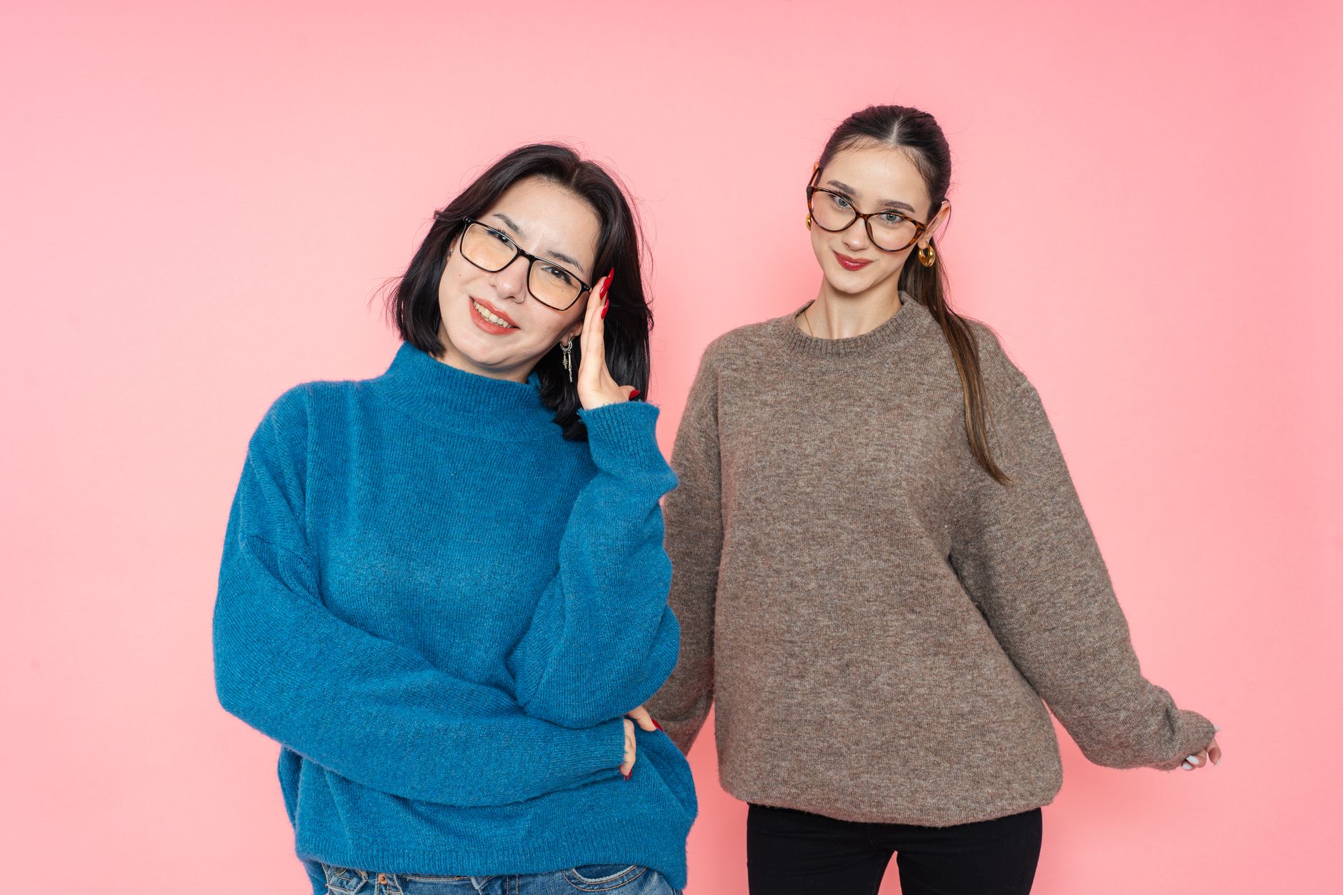 Portrait of two women in sweaters and glasses posing confidently against a pink backdrop.