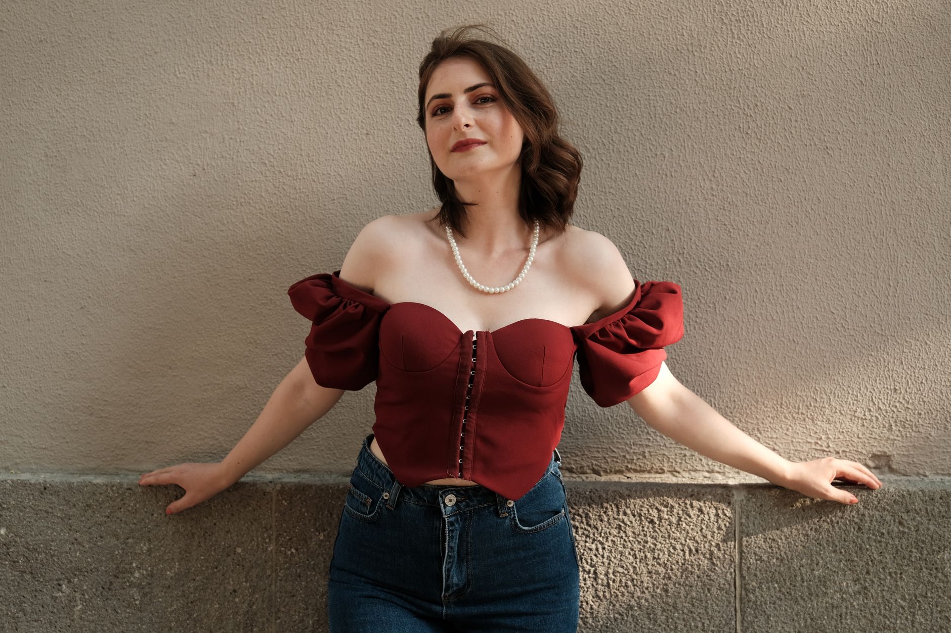 Stylish woman in off-shoulder red blouse poses confidently against a textured wall.