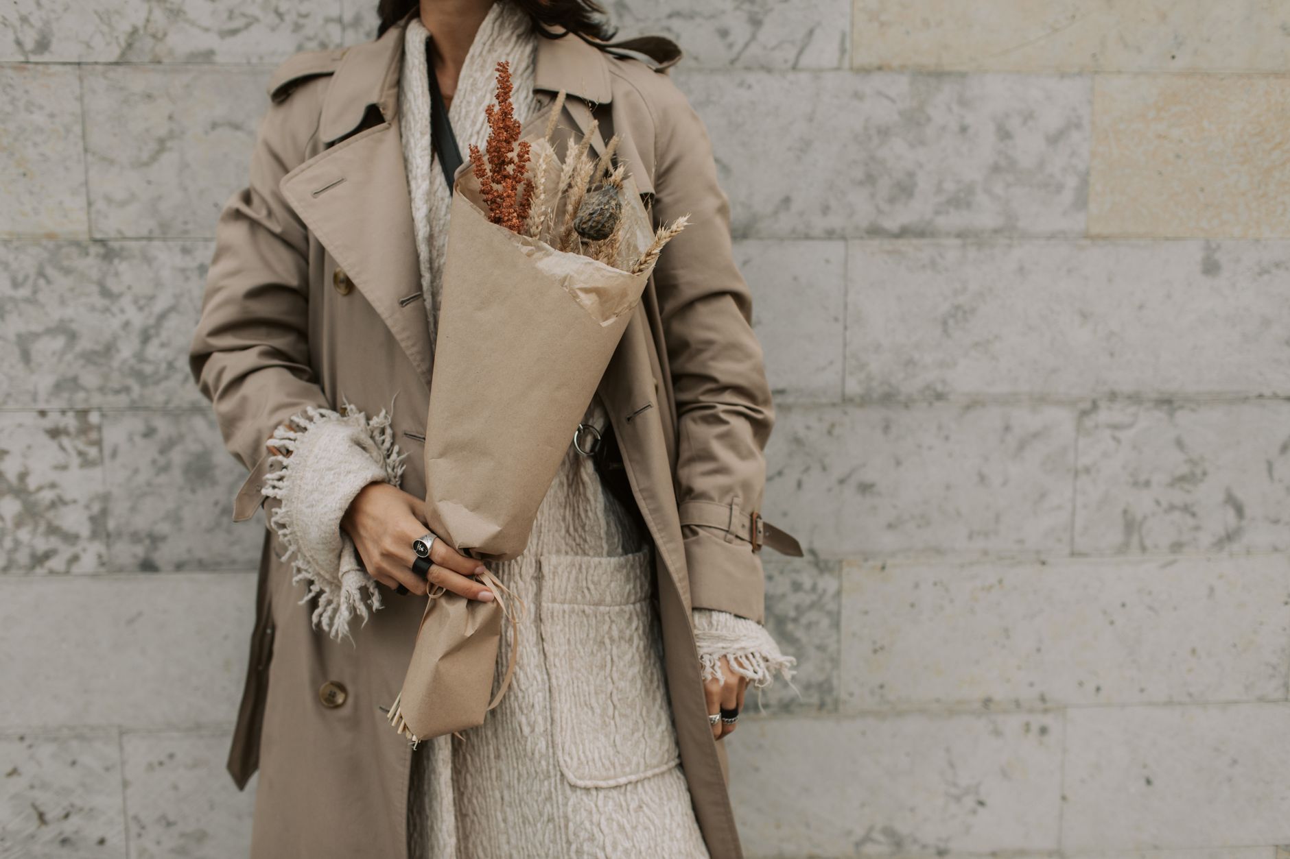 Fashionable woman in a trench coat holding a bouquet of dried autumn flowers against a textured wall.