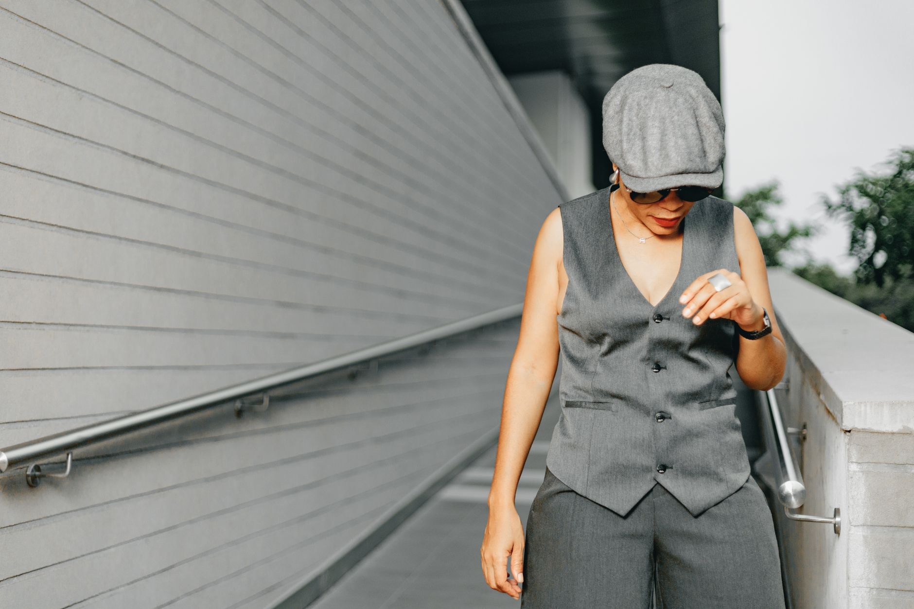 Elegant woman in grey vest and cap poses outdoors on a modern urban pathway.
