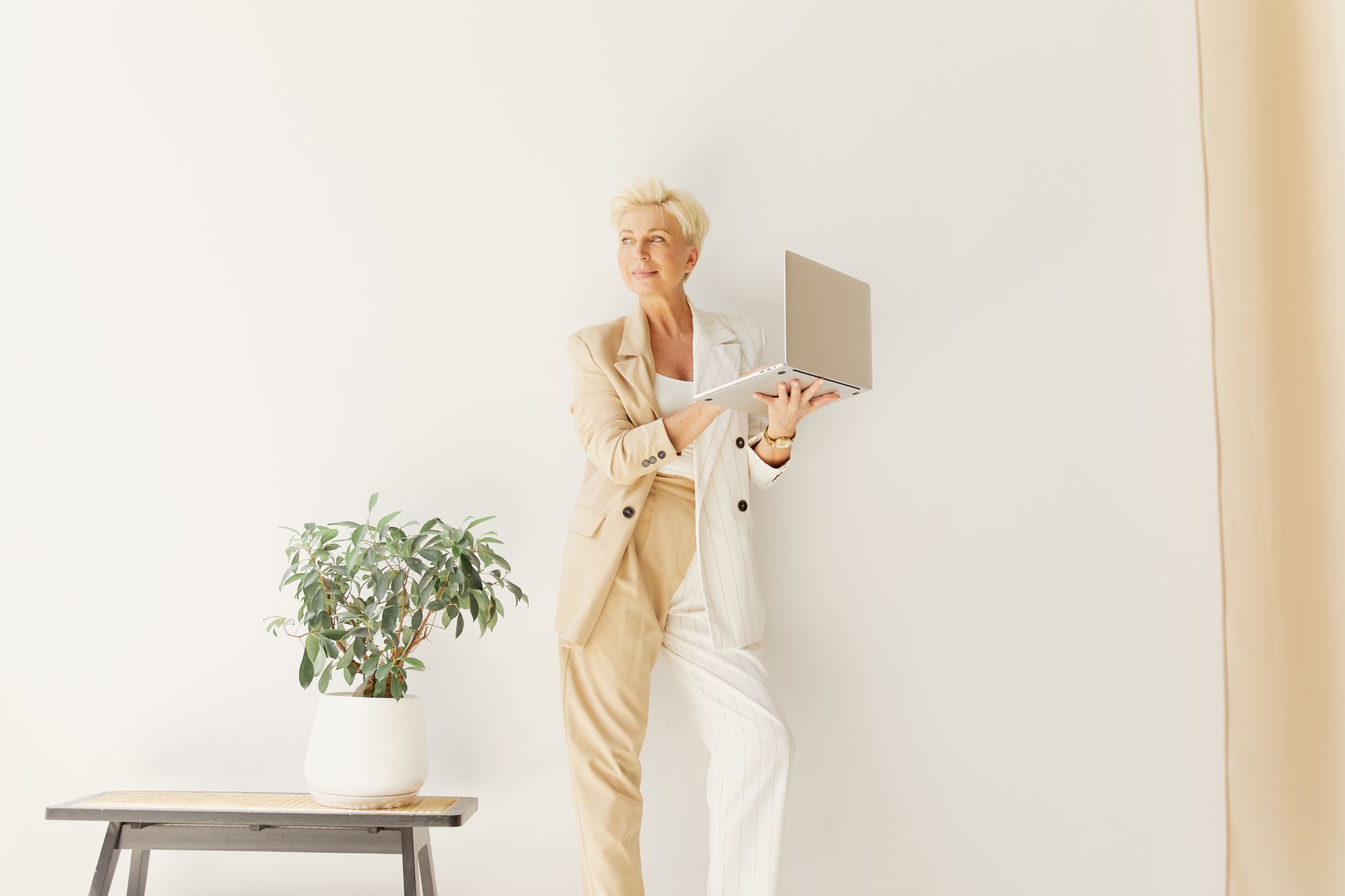 Stylish older woman in a beige suit holding a laptop next to a potted plant indoors.