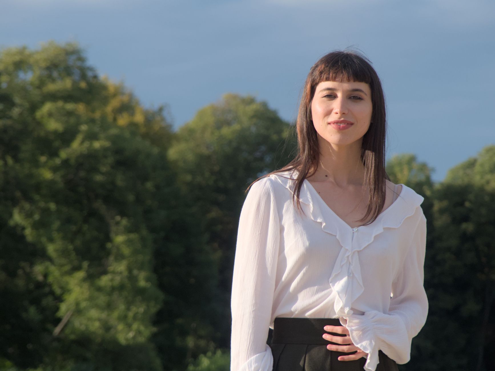 A young woman wearing a ruffled white blouse poses confidently in a park setting.