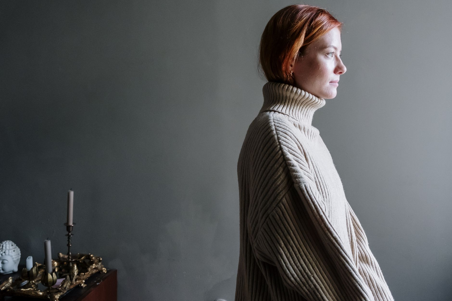A woman in a ribbed turtleneck sweater stands in a minimalist room with soft lighting.