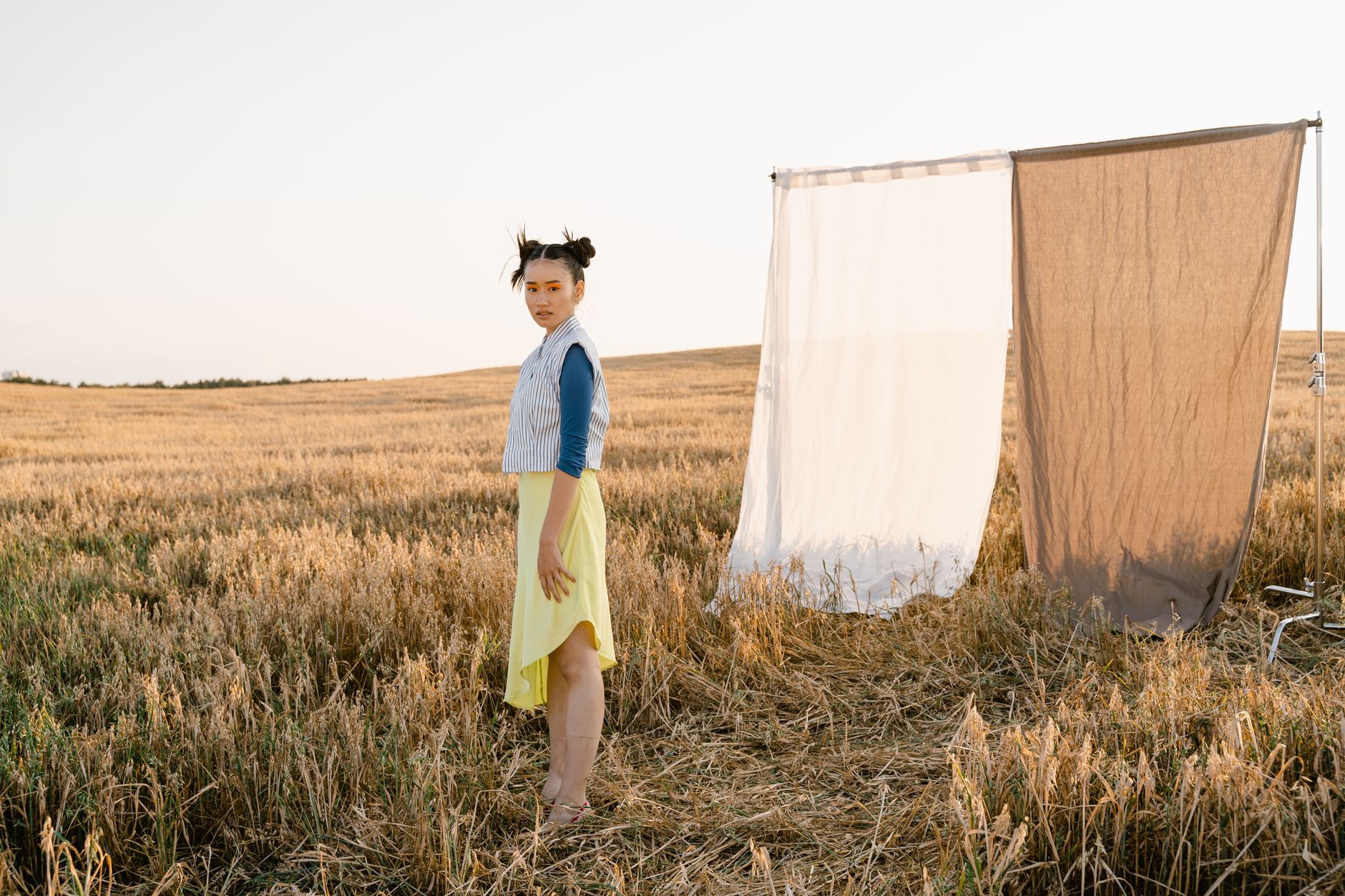 A fashionable woman standing in a field with fabric screens and golden hour lighting, creating a modern outdoor portrait.