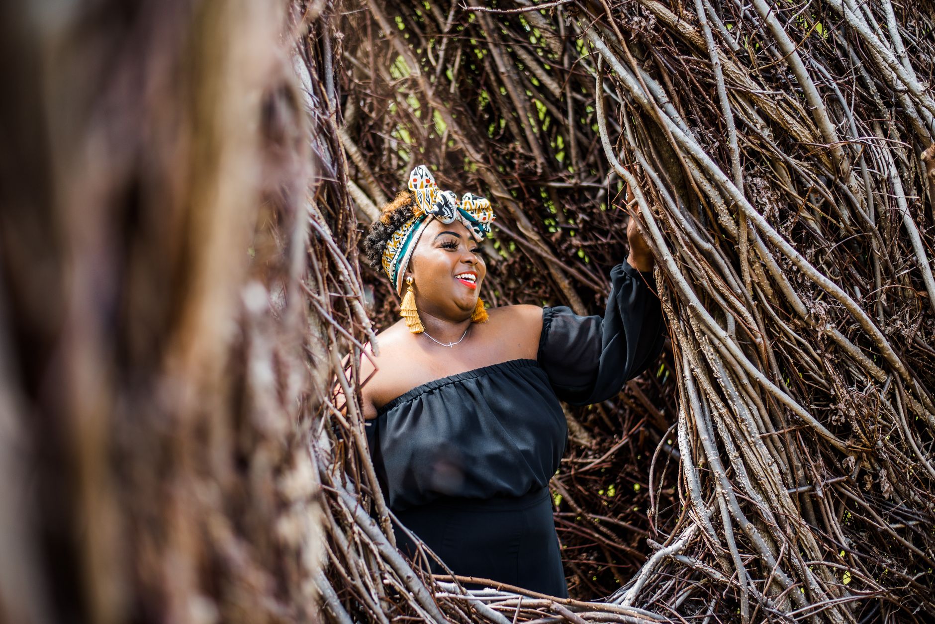 A plus-size woman in a black dress joyfully poses in a natural setting with dry branches.