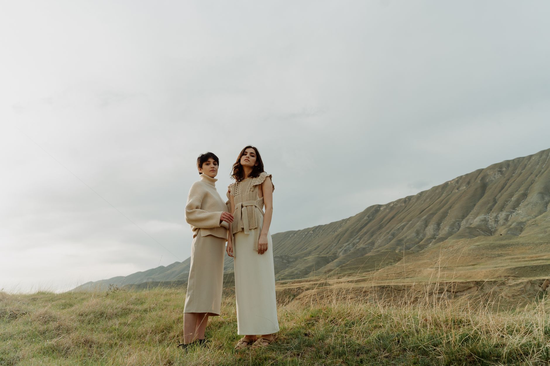 Two women in fashionable beige outfits standing in a serene outdoor landscape.