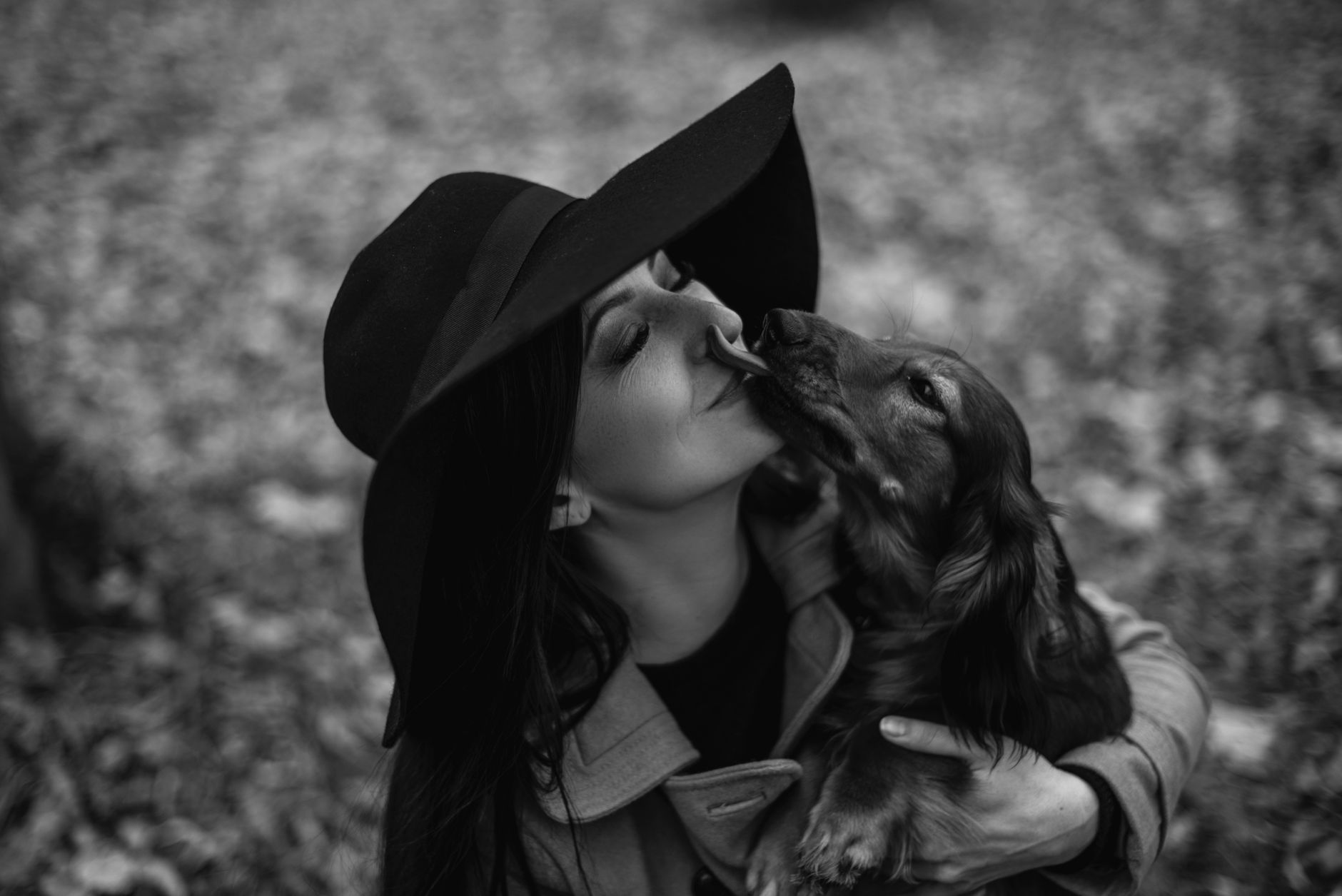 Elegant black and white photo of a woman in a hat with a dachshund outdoors.
