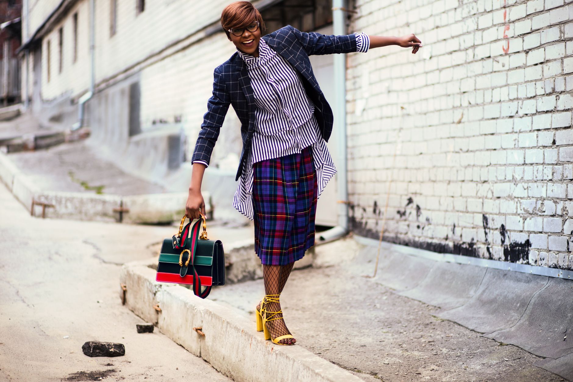 Stylish woman in plaid skirt and blazer playfully balances on outdoor brick street.