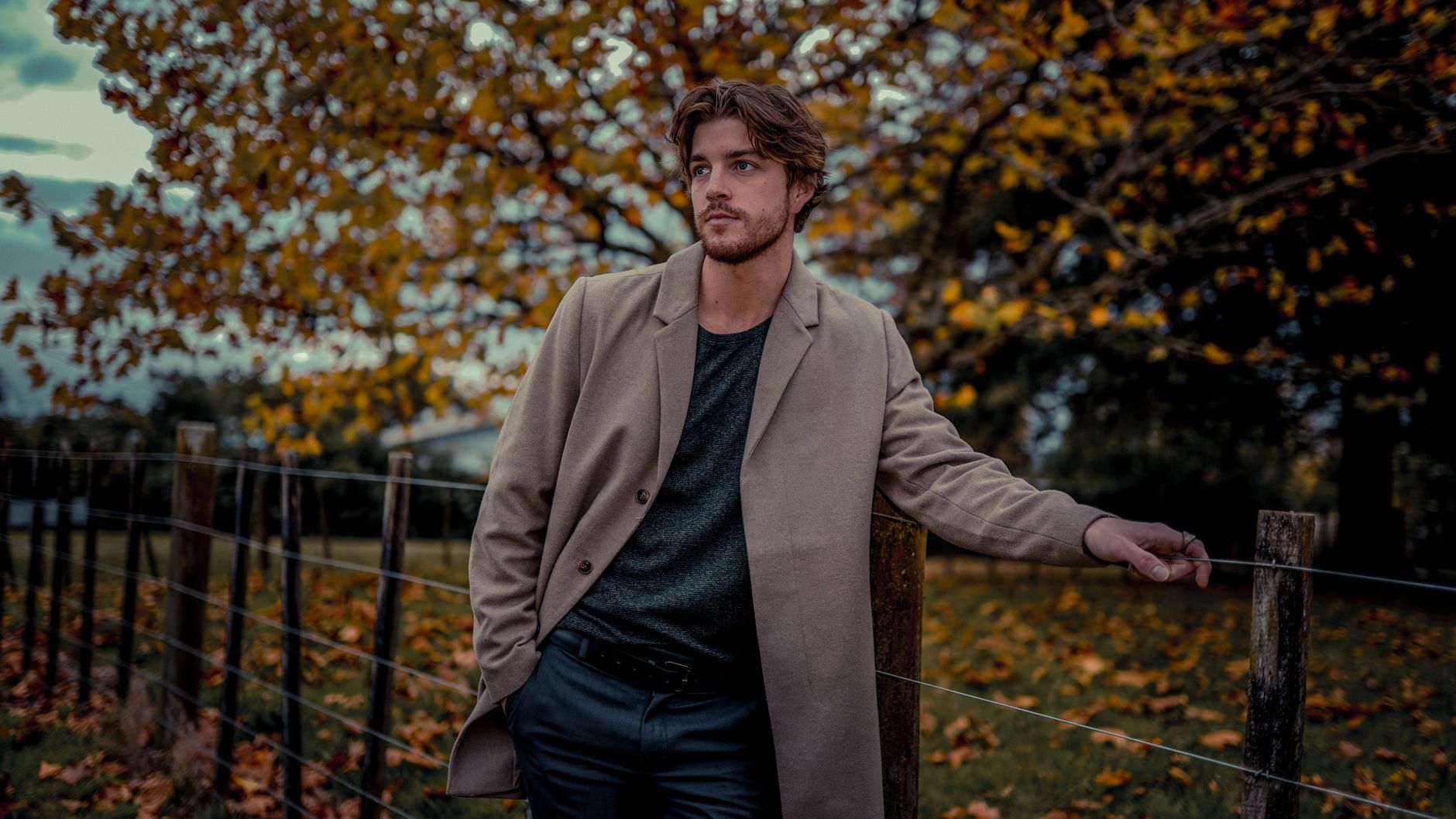 Handsome man in a beige coat posing outdoors with fall foliage background.