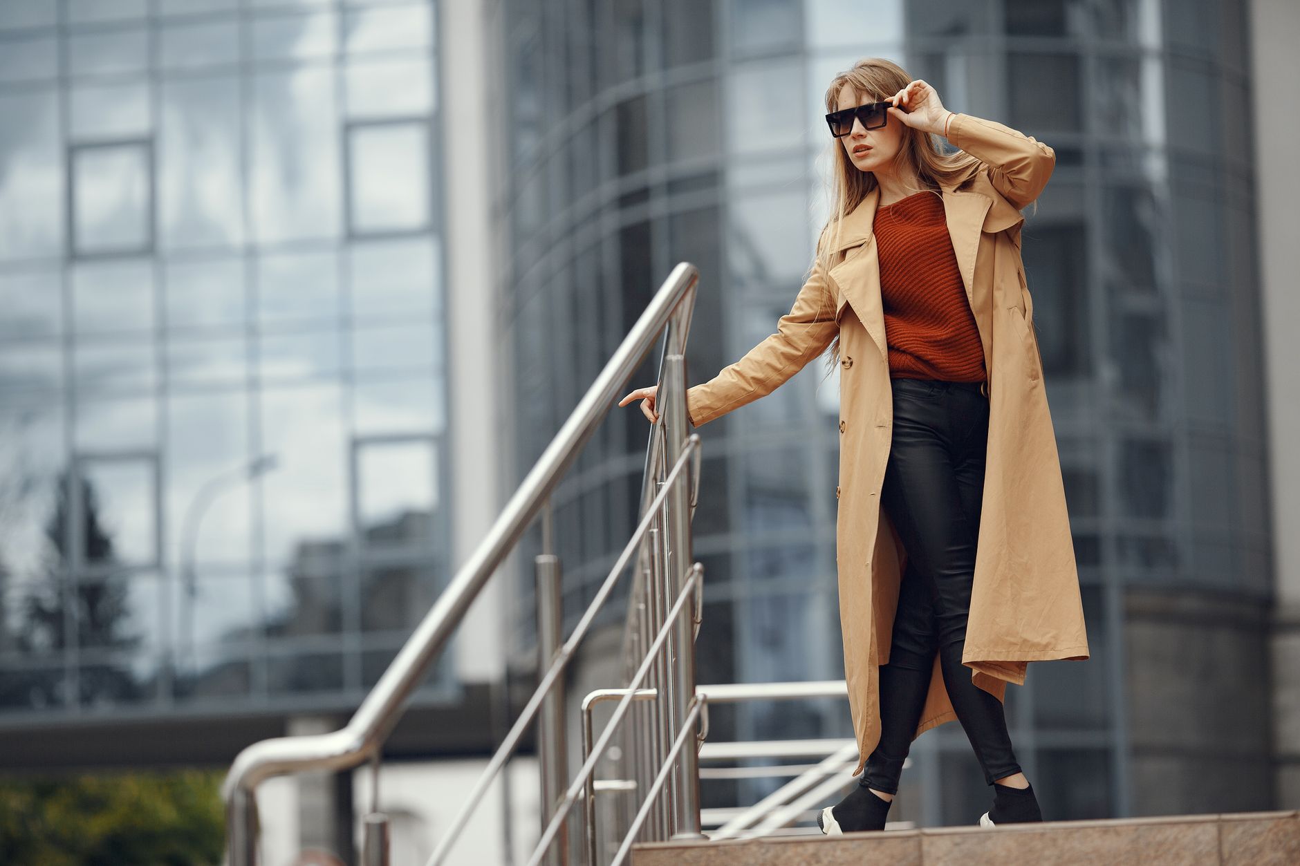 Elegant woman in trench coat adjusting sunglasses on city stairs with office building background.