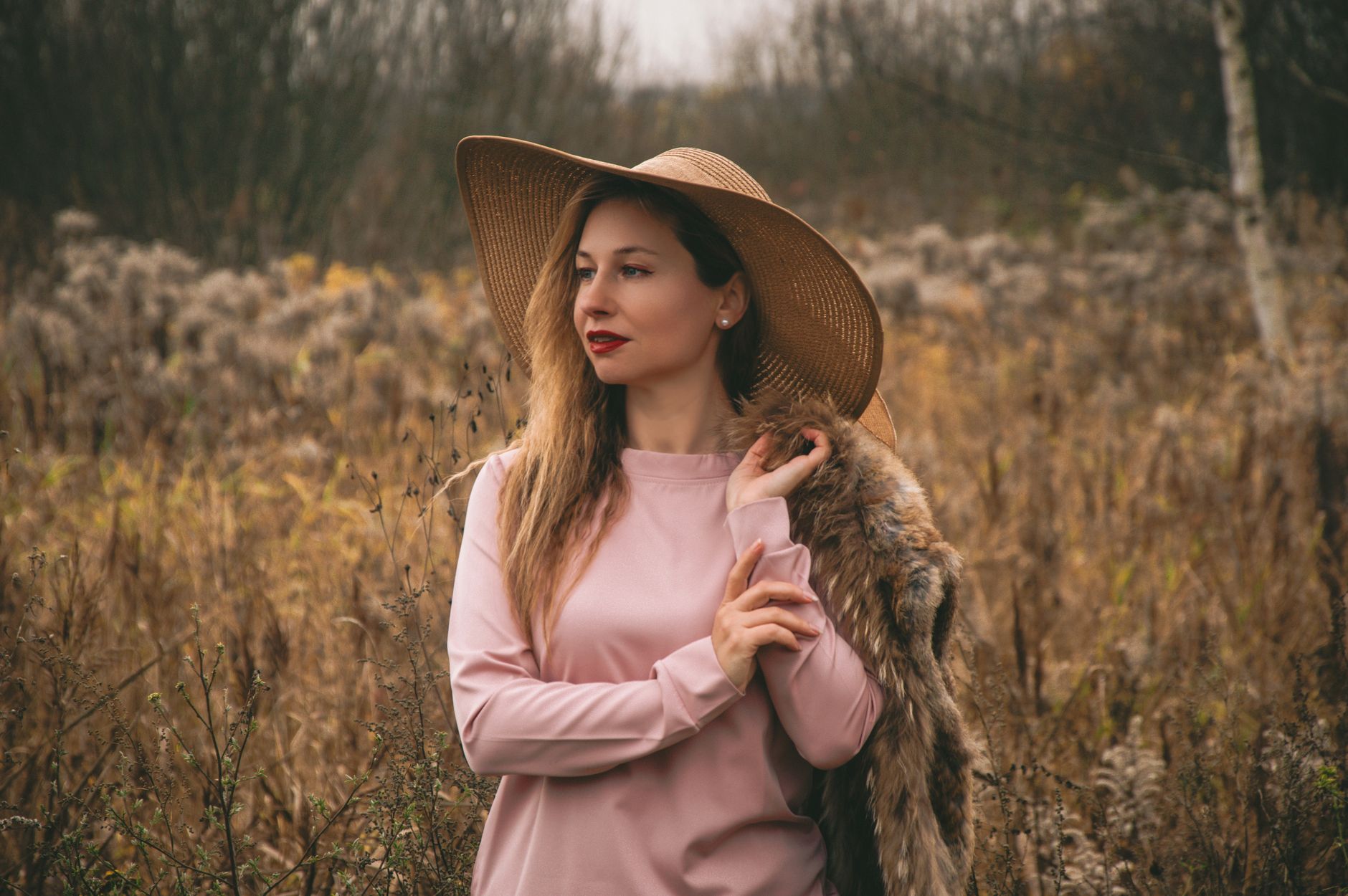 Stylish woman in straw hat and coat posing in a golden autumn field.
