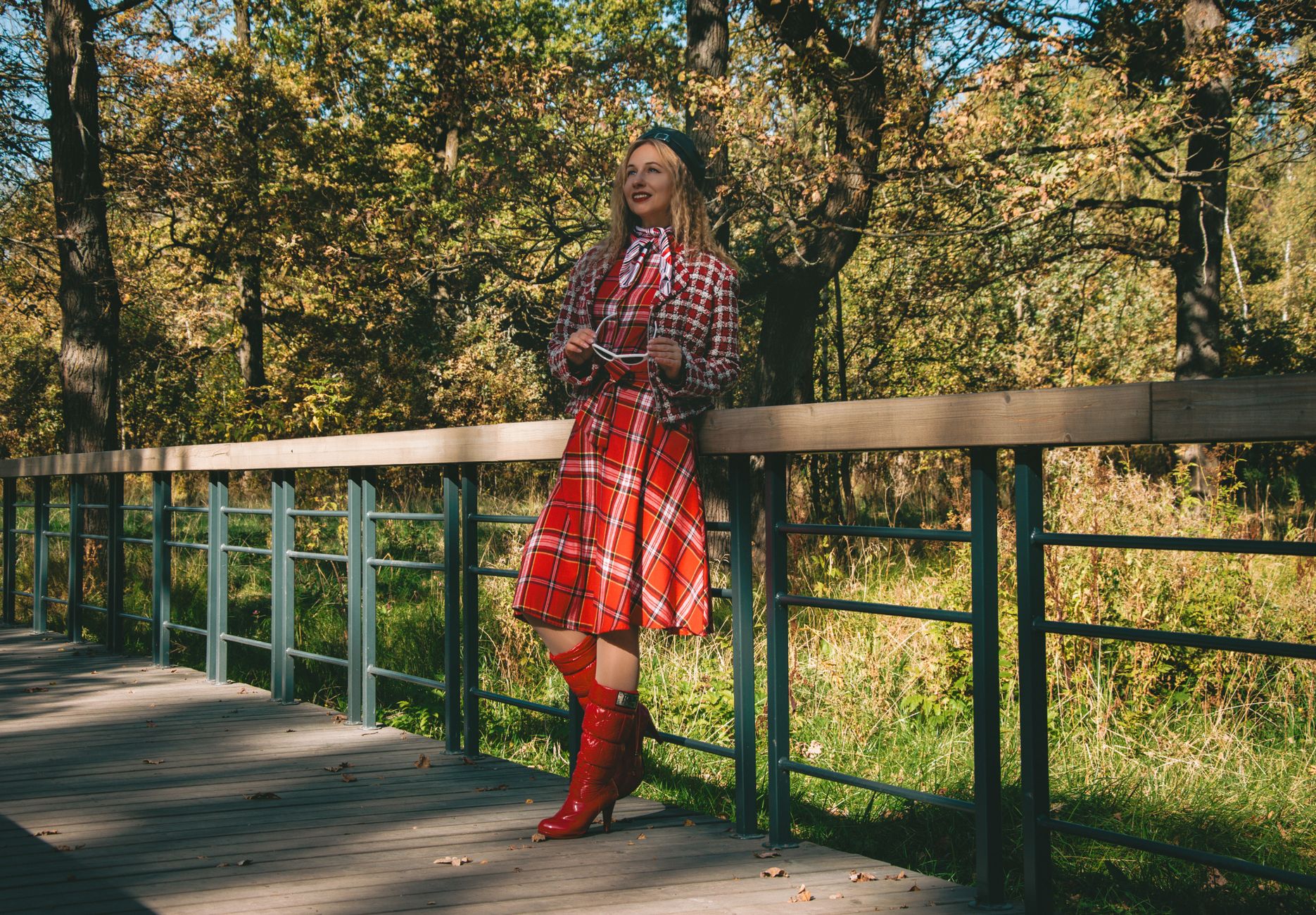 Woman in stylish red plaid outfit standing on a wooden railing surrounded by fall foliage.