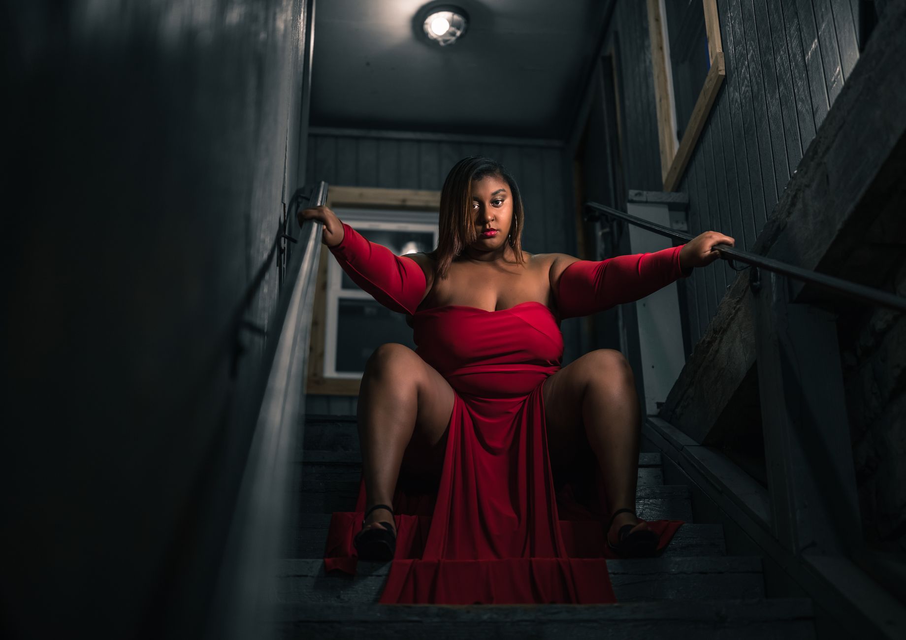 A confident woman in a striking red dress poses on dimly lit stairs, exuding empowerment.
