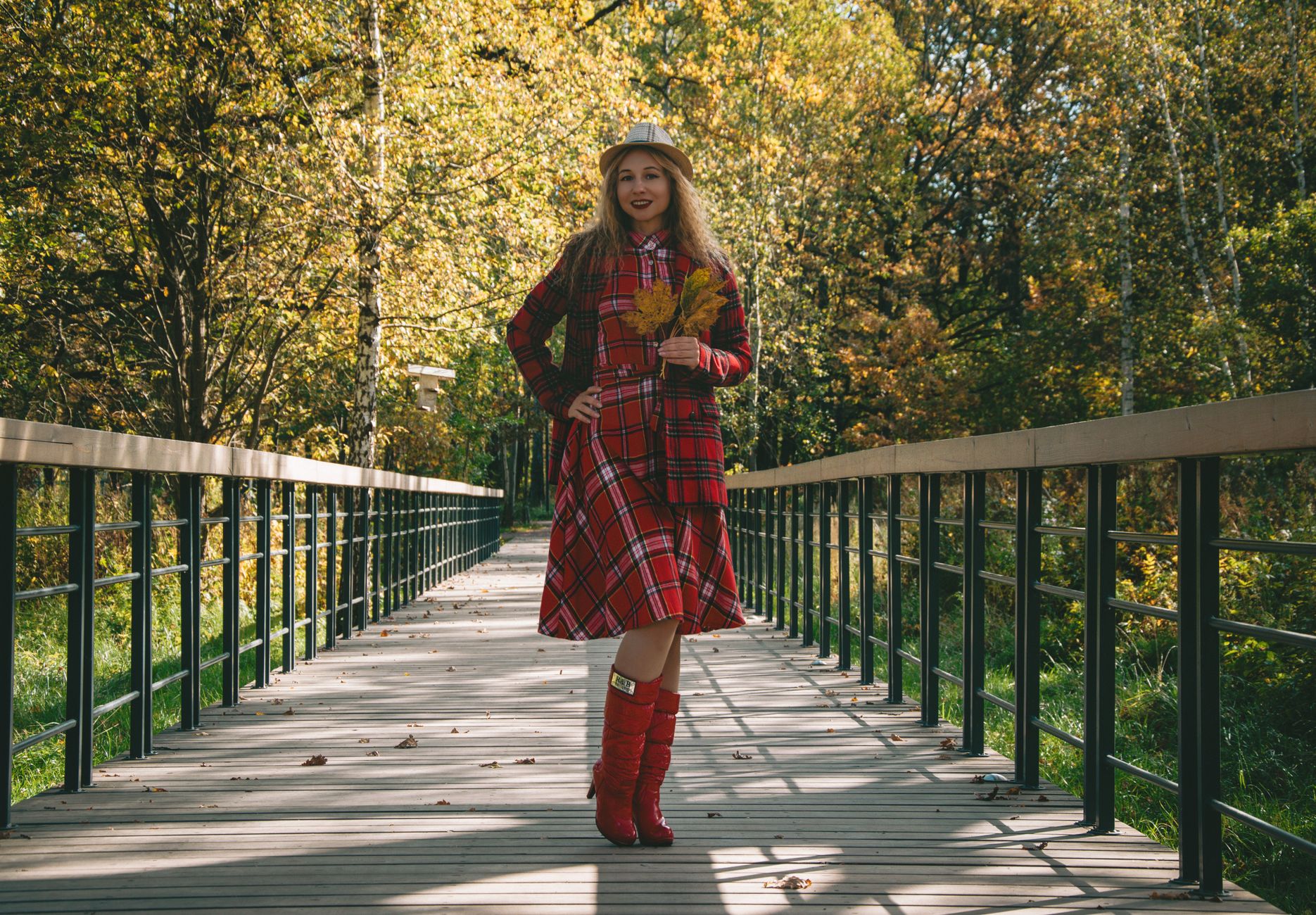 Smiling woman in red tartan dress and boots holding autumn leaves on a bridge
