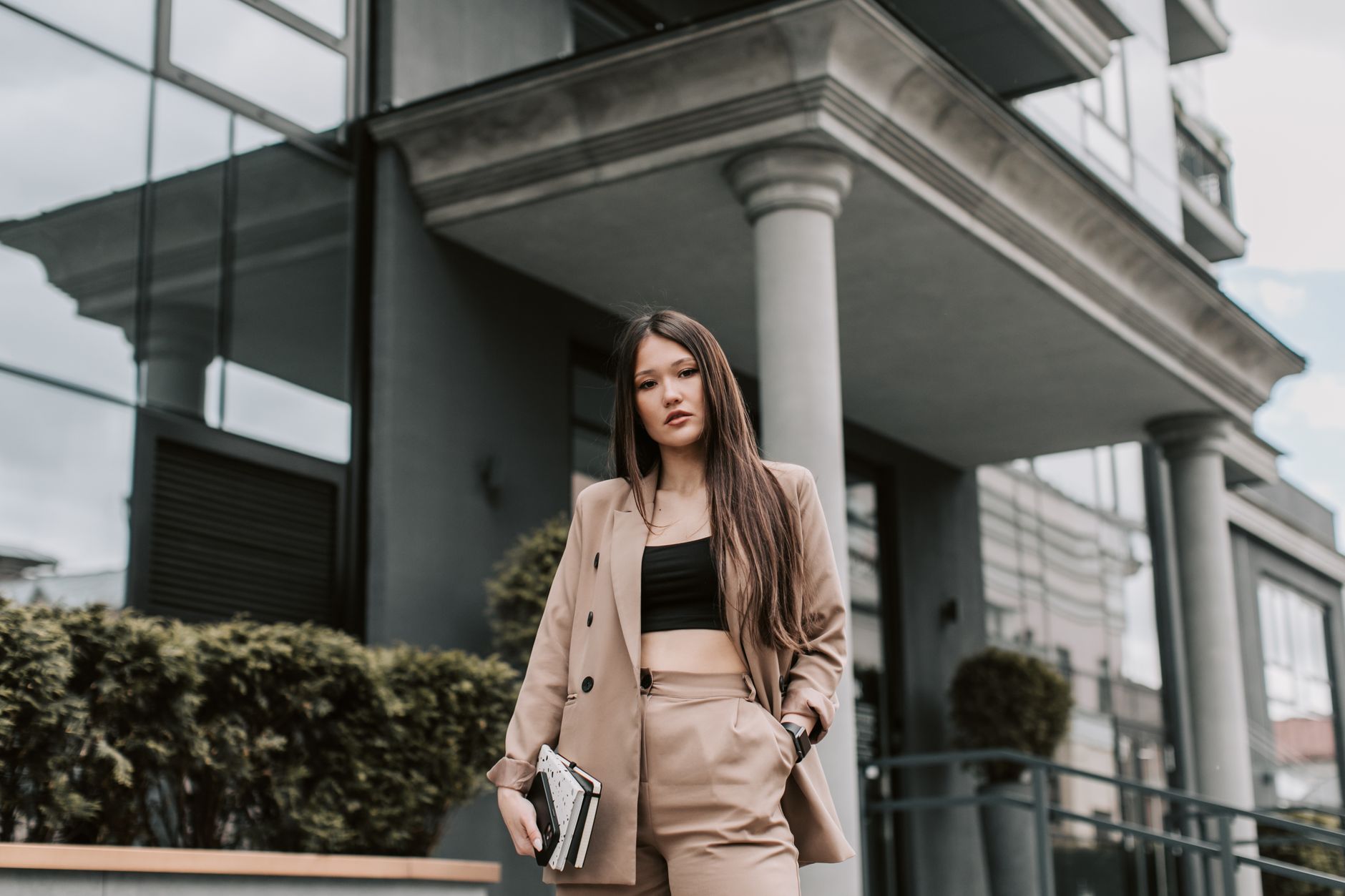 Confident businesswoman in a beige suit standing outdoors, holding notebooks with a modern architecture backdrop.