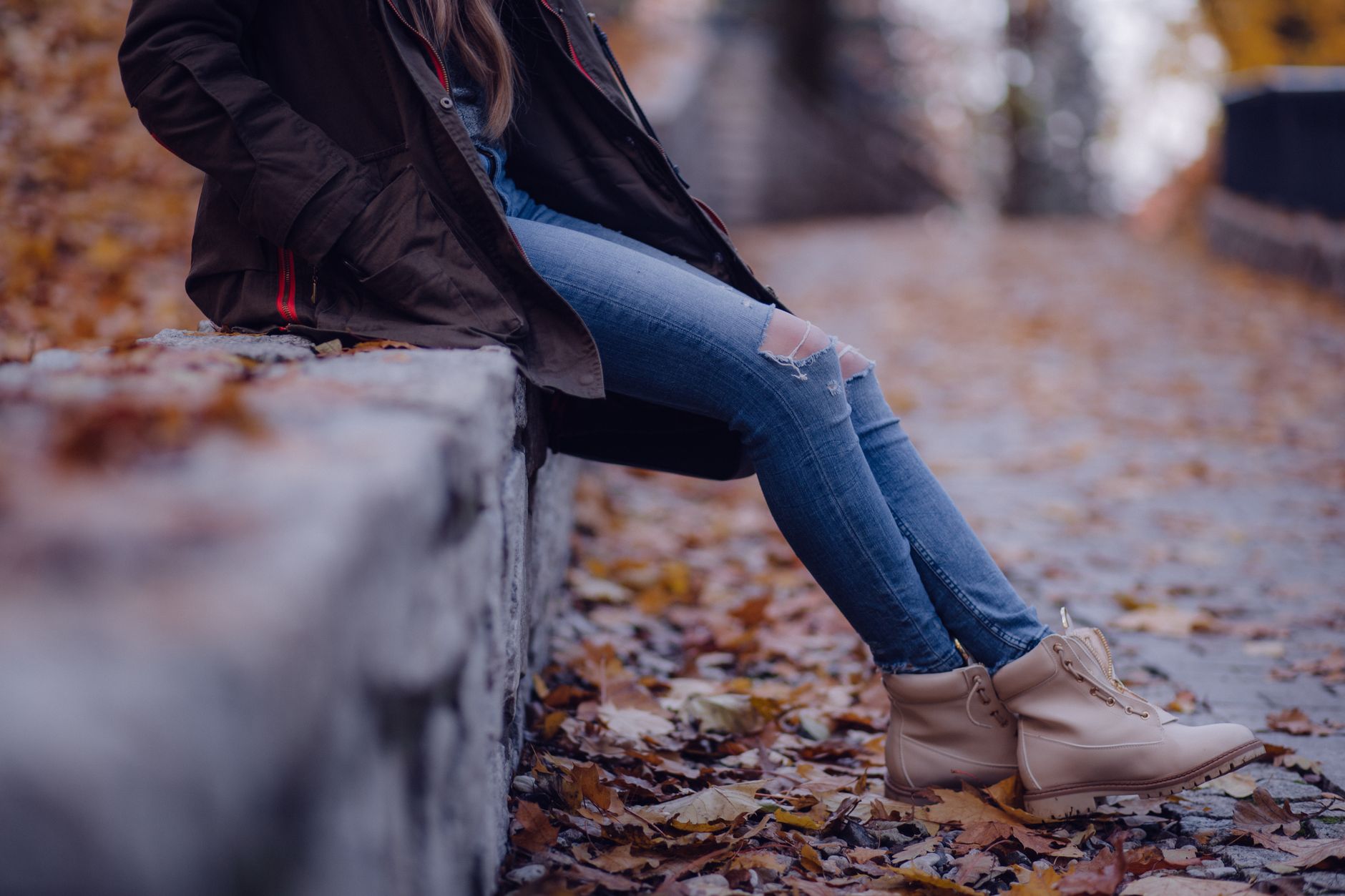 Woman wearing a jacket and boots sits casually on a fall leaf-strewn path in a relaxed mood.